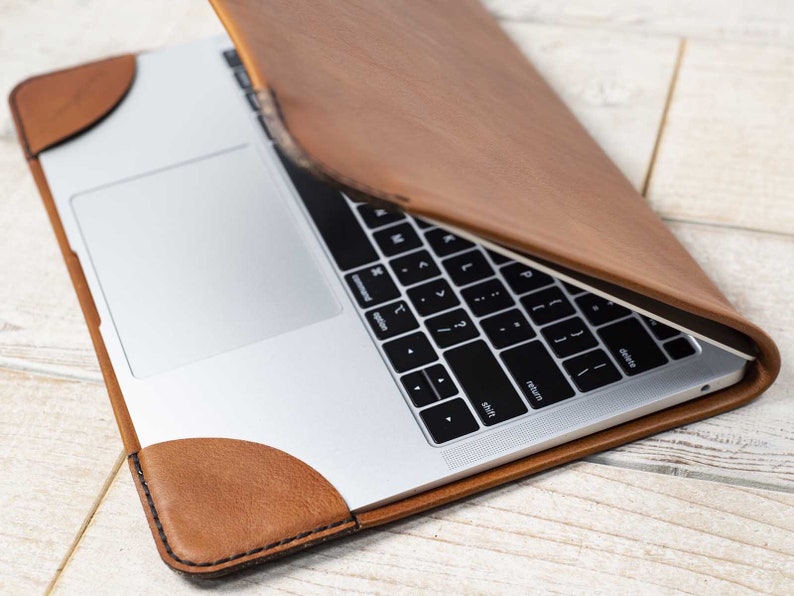 May include: A brown leather laptop sleeve with a laptop inside. The sleeve has rounded corners and a stitched border. The laptop is silver with a black keyboard. The sleeve is open, revealing the laptop's keyboard and trackpad.