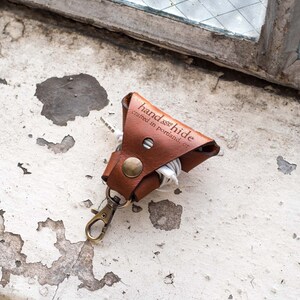May include: A brown leather cord keeper with a metal key ring. The cord keeper has the text "hand hide crafted in portland, or" embossed on it.