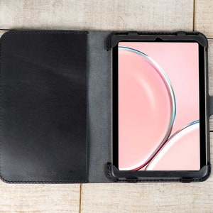 May include: A black leather tablet case with a flap that folds over the screen. The case has a stitched edge and a small metal button on the right side. The tablet screen shows a pink background with abstract shapes.