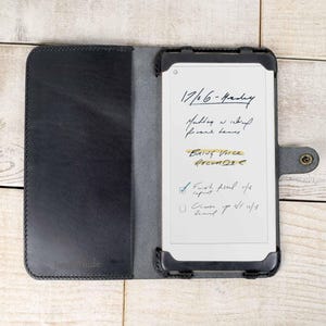 May include: A black leather folio-style case, open to reveal a digital notepad with handwritten notes. The case is open on a light wood surface. The notepad displays a list of tasks and appointments.