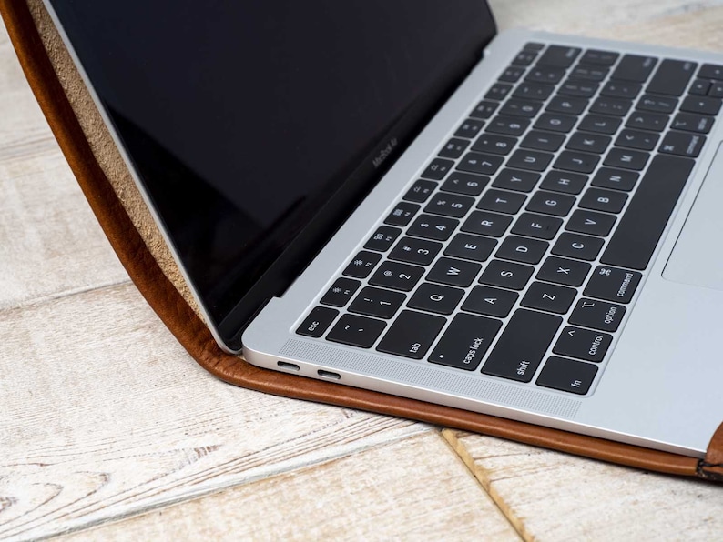 May include: A silver laptop with a black keyboard rests inside a brown leather case. The laptop screen is black and open, displaying a portion of the keyboard. The case has a textured appearance.