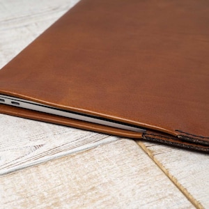 May include: A brown leather laptop sleeve. The sleeve is made of a smooth, textured leather and has a stitched edge. The laptop is partially visible inside the sleeve.