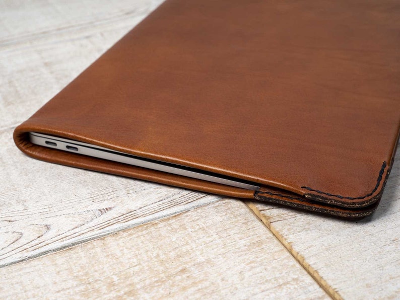 May include: A brown leather laptop sleeve. The sleeve is made of a smooth, textured leather and has a stitched edge. The laptop is partially visible inside the sleeve.