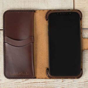 May include: A brown leather phone case with a flap that folds over the screen. The case has a pocket on the outside for cards or cash. The case has the text "hand & hide" embossed on the front.