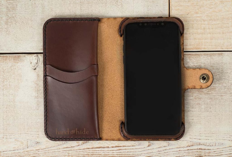 May include: A brown leather phone case with a flap that folds over the screen. The case has a pocket on the outside for cards or cash. The case has the text "hand & hide" embossed on the front.