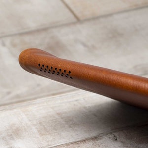 May include: Close-up of a brown leather case, possibly for a tablet or e-reader. The case has a rounded edge with a series of small, evenly spaced holes, likely for a speaker. The leather has a textured appearance.