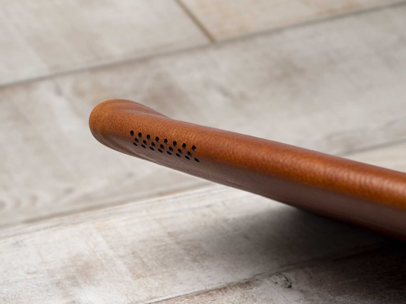 May include: Close-up of a brown leather case, possibly for a tablet or e-reader. The case has a rounded edge with a series of small, evenly spaced holes, likely for a speaker. The leather has a textured appearance.