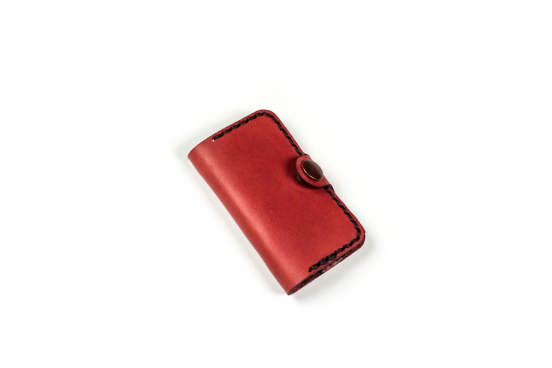May include: A red leather key holder with a button closure. The key holder has black stitching around the edges.