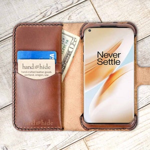 Op de afbeelding: Een bruine leren telefoonhoes met een flap die over het scherm vouwt. De hoes heeft een vak voor kaarten en contant geld. Het telefoonscherm toont de woorden "Never Settle".