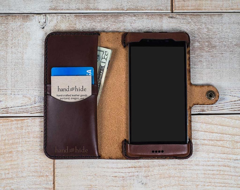 May include: A brown leather phone case with a card slot and a flap that closes with a snap. The case has the text "hand hide" and "hand-crafted leather goods portland, oregon, usa" printed on it.