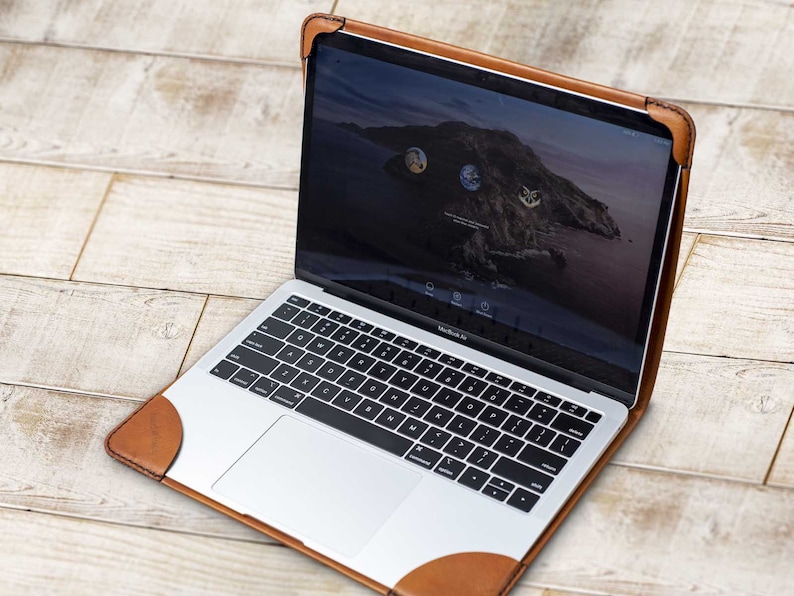 May include: A silver MacBook Air laptop in a brown leather case. The case has reinforced corners and a stitched design. The laptop screen displays a scenic background with icons. The keyboard is black and white.