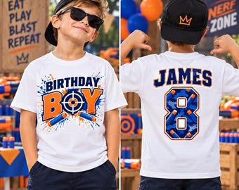 May include: A white short-sleeve t-shirt featuring a front graphic with the text "BIRTHDAY BOY" in blue and orange with a target icon and foam dart imagery. The back displays the name "JAMES" and the number "8" in a blue and orange camo pattern.
