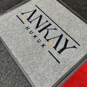 Puede incluir: Una alfombrilla rectangular gris con superficie texturizada que presenta el texto azul oscuro ANKAY HUKUK impreso en una fuente serif negrita. El texto está flanqueado por dos líneas horizontales y dos pequeños acentos de diamante amarillo.