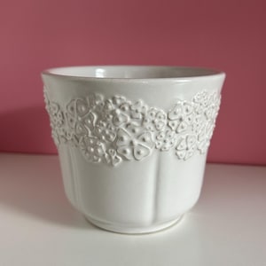 Peut inclure: Un pot de fleurs en céramique blanche avec un motif floral texturé sur le dessus. Le pot a une forme arrondie et des rainures verticales. Le fond est de couleur rose uni.