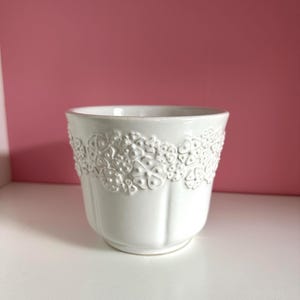 May include: A white ceramic plant pot with a textured floral pattern around the rim. The pot has a rounded shape and vertical grooves on the lower section. It is on a white surface against a pink backdrop.