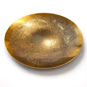 Pode incluir: Um prato decorativo redondo, dourado, com um design floral em relevo ornamentado. O prato tem uma superfície brilhante e reflexiva e uma borda ligeiramente elevada. O padrão intrincado cobre a maior parte da superfície do prato.