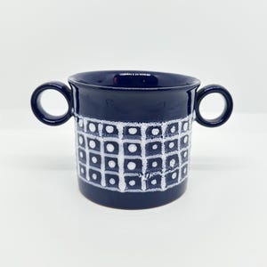 May include: A dark blue ceramic mug featuring two circular handles on opposite sides. The exterior surface is decorated with a white grid pattern containing small white dots within each square, creating a textured and repeating geometric design.
