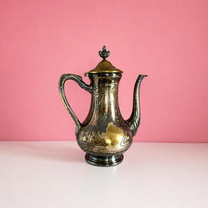 Peut inclure: Une cafetière antique argentée avec une poignée et un bec décoratifs. La cafetière a un couvercle doré et un motif floral détaillé. La cafetière est présentée sur une surface blanche sur fond rose.