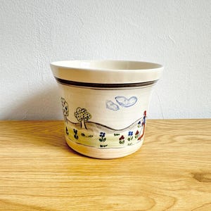 Può includere: Un vaso da fiori in ceramica color crema con un paesaggio dipinto a mano raffigurante alberi verdi, nuvole blu, fiori colorati e una figura con un vestito rosso. Il vaso presenta un bordo ampio e svasato con una fascia orizzontale marrone.