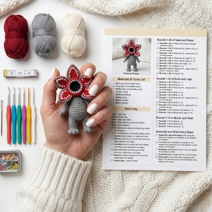 Könnte beinhalten: Handgefertigte Demogorgon-Puppe aus Häkelarbeit, gehalten in einer Hand mit weißem Nagellack, umgeben von Häkelzubehör. Enthält rote, graue und cremefarbene Wolle, Häkelnadeln und ein Schnittmuster.