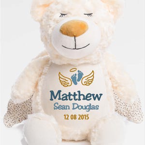 Puede incluir: Oso de peluche color crema con detalles bordados. Presenta ojos cerrados, una nariz marr&oacute;n y una boca sonriente. El pecho del oso muestra alas doradas, huellas azules y el nombre "Matthew Sean Douglas" con la fecha "12 08 2015".