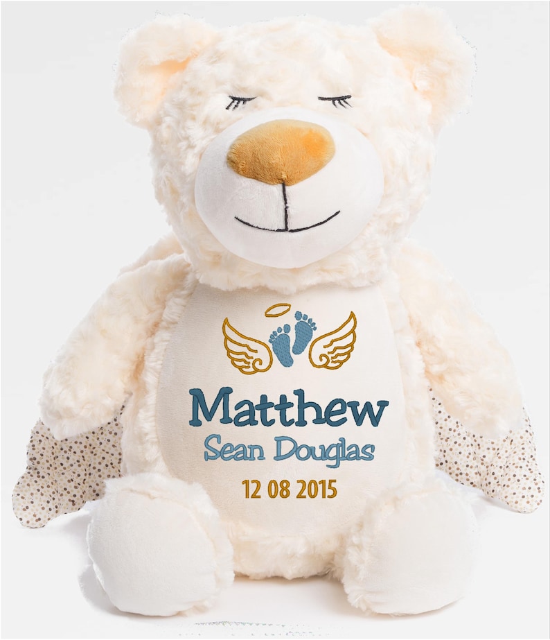Puede incluir: Oso de peluche color crema con detalles bordados. Presenta ojos cerrados, una nariz marr&oacute;n y una boca sonriente. El pecho del oso muestra alas doradas, huellas azules y el nombre "Matthew Sean Douglas" con la fecha "12 08 2015".