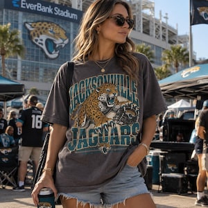 May include: A charcoal gray short-sleeve t-shirt featuring a graphic of a roaring jaguar, a football helmet, and the text Jacksonville Jaguars in teal and gold lettering. The shirt has a relaxed fit and is paired with light blue denim shorts.