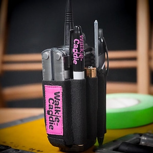 Może przedstawiać: Czarny pokrowiec na krótkofalówkę z różową etykietą z napisem "Walkie Caddie". Pokrowiec mieści wielofunkcyjne narzędzie, czarny marker z napisem "Walkie Caddie" i inne akcesoria. Pokrowiec jest przymocowany do krótkofalówki.