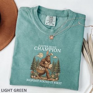 May include: Light green Comfort Colors t-shirt with the text "EASTER HUNT CHAMPION" and "BIGFOOT FOUND IT FIRST". The graphic depicts Bigfoot with bunny ears carrying a basket of Easter eggs. A silver necklace is draped across the shirt.