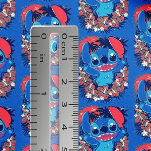 May include: Blue fabric featuring a repeating pattern of a cartoon character with large eyes, surrounded by red flowers and palm trees. A ruler shows measurements in inches. The character is blue with red ears and a red mouth.