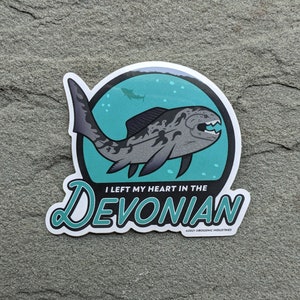 May include: A sticker depicting a grey and black prehistoric fish with sharp teeth, set against a teal background with bubbles and a smaller fish. The text reads "I LEFT MY HEART IN THE DEVONIAN."