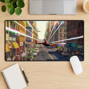 May include: A black rectangular mousepad featuring a vibrant illustration of a superhero in a city setting. The superhero is in mid-air, with buildings and streets in the background. The mousepad is on a wooden desk with a laptop, plant, coffee cup, notepad, and mouse.