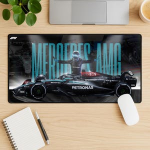 May include: Black rectangular mousepad featuring a Formula 1 race car with the text "MERCEDES AMG" in teal. A driver in a helmet is in the center with arms outstretched. The mousepad has a black background with the Petronas logo.