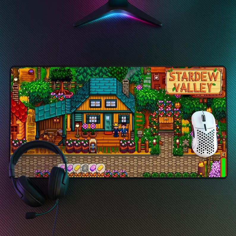 May include: A rectangular Stardew Valley mousepad featuring a pixelated scene of a house, characters, and the game's logo. A white mouse and black headset are on the pad. The pad has a black textured surface.