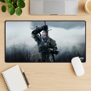 Könnte beinhalten: Schwarzes rechteckiges Mauspad mit einem Bild eines Mannes in Rüstung, der ein Schwert hält, vor einem nebligen Waldhintergrund. Das Mauspad hat den Text "THE WITCHER" unten rechts. Eine weiße Computermaus befindet sich auf der rechten Seite.