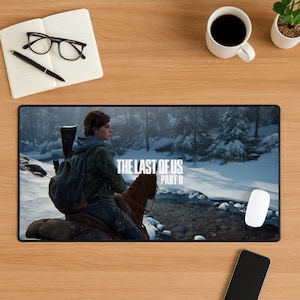 May include: Black rectangular mousepad featuring a scene from "The Last of Us Part II" video game. The image shows a character riding a horse in a snowy forest. Includes the game's title in white text. A white mouse and a smartphone are also visible.