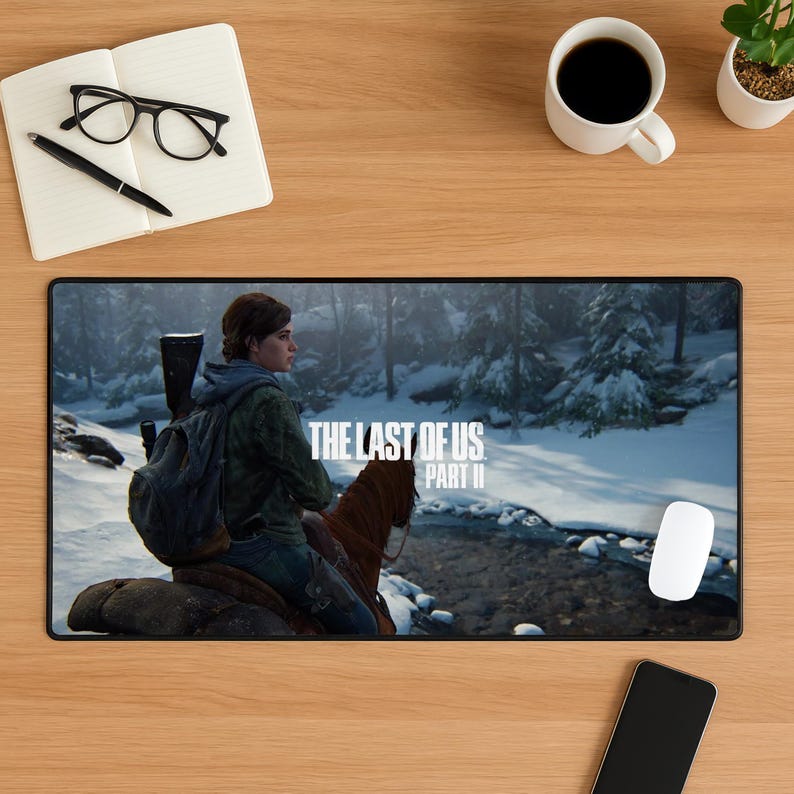 May include: Black rectangular mousepad featuring a scene from "The Last of Us Part II" video game. The image shows a character riding a horse in a snowy forest. Includes the game's title in white text. A white mouse and a smartphone are also visible.