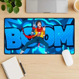 Puede incluir: Alfombrilla de ratón rectangular negra con un personaje de dibujos animados con una chaqueta amarilla y vaqueros azules, haciendo un gesto de rock and roll. El fondo es azul brillante con la palabra "BOM". Se ve un ratón blanco y un bloc de notas.