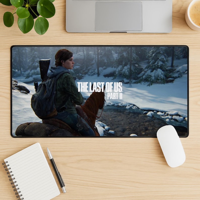 May include: Black rectangular mousepad featuring a scene from "The Last of Us Part II" video game. The image shows a woman on horseback in a snowy forest. The game's title is printed in white.