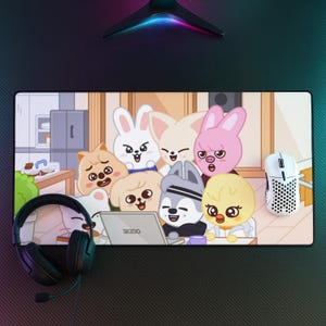 以下が含まれることがあります： ラップトップの周りに集まった動物のキャラクターの漫画イラストが描かれた長方形のデスクマット。黒いヘッドセットと白いマウスがマットの上に配置されています。ラップトップには「SK230」というテキストが表示されています。
