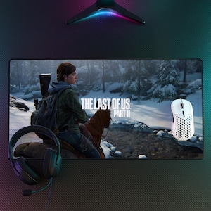 May include: A gaming mouse pad featuring the game "The Last of Us Part II" with a scene of a woman on horseback. A white gaming mouse and black headset are also visible. The pad is rectangular and has a black background.