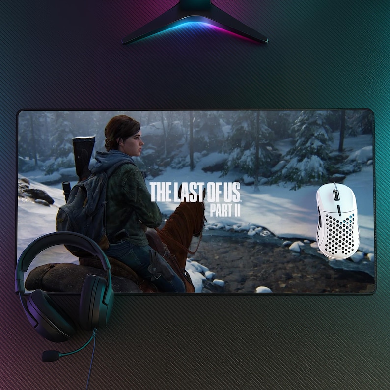 May include: A gaming mouse pad featuring the game "The Last of Us Part II" with a scene of a woman on horseback. A white gaming mouse and black headset are also visible. The pad is rectangular and has a black background.