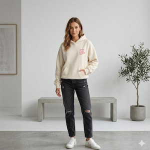 May include: A cream-colored hoodie with a front pocket and a pink embroidered design on the chest. The model is wearing black distressed jeans and white sneakers. A bench and a potted plant are in the background.