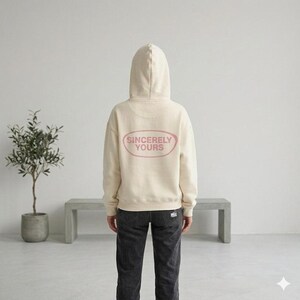May include: Cream-colored hoodie with a hood and the text "Sincerely Yours" in a pink oval on the back. The hoodie is paired with dark gray jeans. The image is taken indoors.