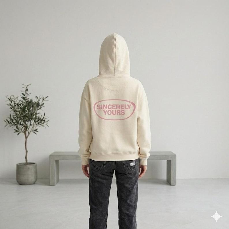 May include: Cream-colored hoodie with a hood and the text "Sincerely Yours" in a pink oval on the back. The hoodie is paired with dark gray jeans. The image is taken indoors.