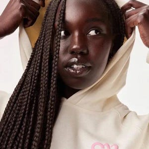 May include: A person wearing a cream-colored hoodie with a pink logo on the chest. The hoodie's hood is up, and the person's dark hair is styled in braids. The person is looking off to the side.