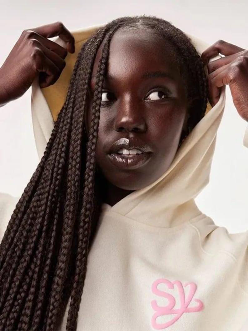 May include: A person wearing a cream-colored hoodie with a pink logo on the chest. The hoodie's hood is up, and the person's dark hair is styled in braids. The person is looking off to the side.