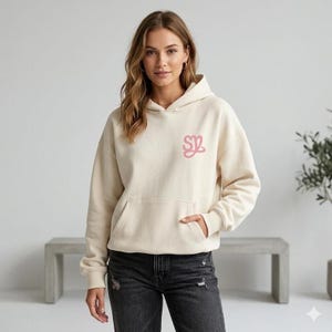 May include: Cream-colored hoodie with a kangaroo pocket and a drawstring hood. The hoodie features a pink embroidered logo on the chest. The person in the image is wearing black denim jeans.