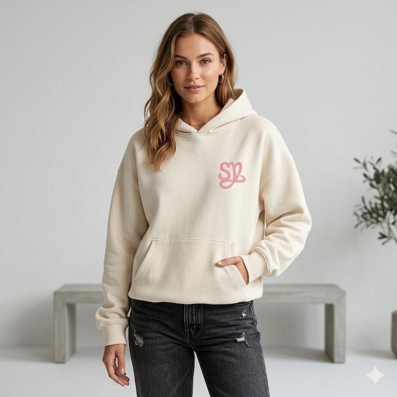 May include: Cream-colored hoodie with a kangaroo pocket and a drawstring hood. The hoodie features a pink embroidered logo on the chest. The person in the image is wearing black denim jeans.
