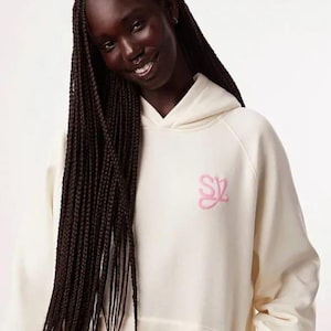 May include: Cream-colored hoodie with a front pocket and a hood. The hoodie features a pink embroidered logo on the chest. The model is wearing long, dark braids. The hoodie is made of a soft, comfortable material.